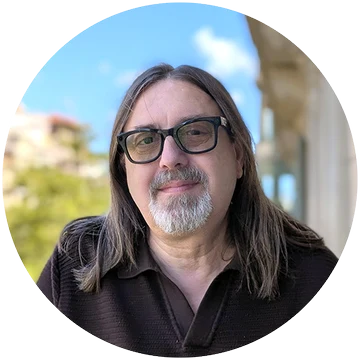 Retrato de Joan Vilariño: melena larga, barba canosa y gafas de pasta, con cielo azul de fondo
